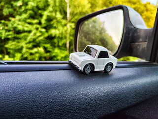 Small white toy car on background of side mirror of a real big caron and nature landscape. Concept of travel and adventure by car. Freedom of movement on your own transport. Blur and partial focus