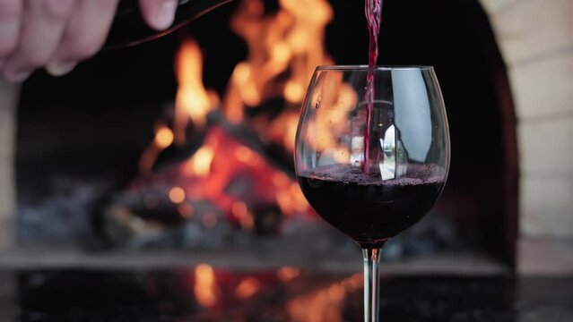 Male hand pouring red wine from bottle to glass goblet at rustic fireplace romantic relaxing slowmo