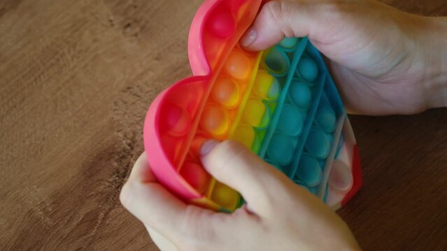 Colorful rainbow antistress sensory toy fidget push pop it on human hand. Young woman plays simple dimple. Rubber multicolored heart in girl's hands, Trending things popular among children and adults.