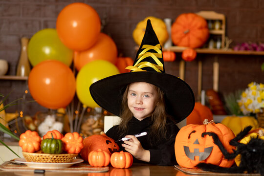 halloween, a child girl in a witch costume with pumpkins and a big spider in a dark kitchen paints a pumpkin for the Halloween holiday, smile, rejoice