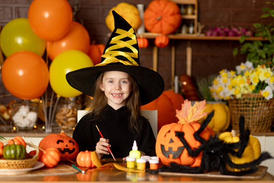 halloween, a child girl in a witch costume with pumpkins and a big spider in a dark kitchen paints a pumpkin for the Halloween holiday, smile, rejoice