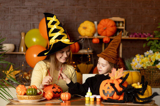 halloween, mom and child daughter in a witch costume with pumpkins and a big spider in a dark kitchen paint a festive pumpkin with paints drawing a muzzle, smile, rejoice