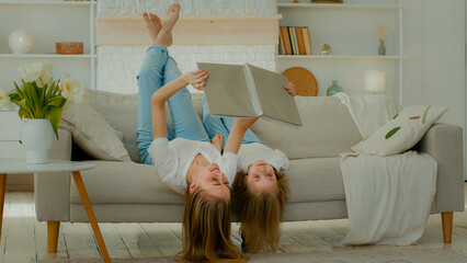 Cheerful Caucasian mother lying upside down on comfortable sofa with little child girl kid daughter...