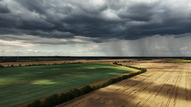 Dramatic dark sky and big rain clouds over the fields. Heavy rain in the distance. Wind turbines at the edge of the picture. Flight along cultivated fields. High voltage power line cuts through landsc