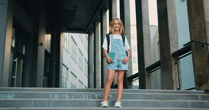 Cheerful girl hugging books posing with backpack near school building outdoor, smiling to camera. Modern education, college tuition and grants, studentship and study abroad concept