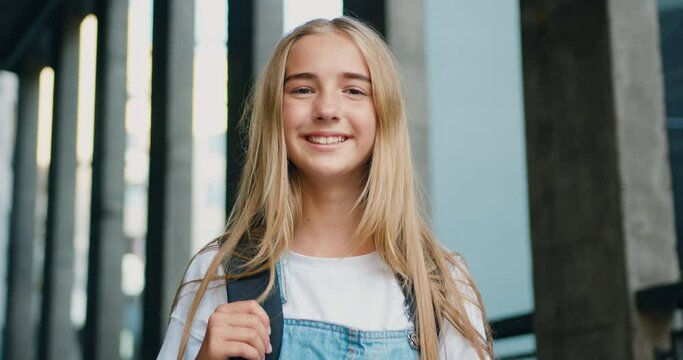 Portrait happy preteen girl holding book and looking at camera outdoors. Portrait of schoolgirl with backpack smiling at camera outside school building.