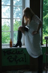 A woman in a white dress puts her foot in a black boot on the windowsill. A bottle of beer on the windowsill. Papers on the floor. On the wall there is an inscription 