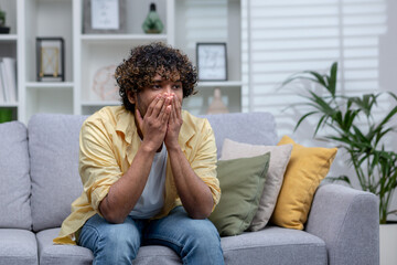 Sad depressed man sitting alone at home on sofa in living room, desperate indian man.