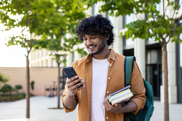 Young smiling Indian male student came to study, standing outside campus, university and using phone. He was holding books and a backpack © Tetiana