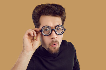 Portrait of funny alert and curious man looking at you through glasses with enlarged lenses. Close up of young Caucasian man isolated on beige background. Concept of own vision, suspicion and humor.