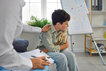 Therapist trying to help unhappy, resentful kid who has behaviour issues. Portrait of sad caucasian boy hugging wooden man figurine to relieve stress in psychologists office. Psychotherapist interior. - obrazy, fototapety, plakaty