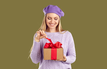 Happy young woman opening gift box with red bow xmas present. Portrait of beautiful smiling girl in lilac beret untying ribbon on gift package celebrating Birthday, Christmas or Valentines Day