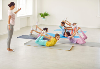 Sporty children do gymnastic exercises. Fit kids practice yoga poses with professional trainer. Group of flexible sportive girls learning to do Dhanurasana bow position on gym floor at sports school