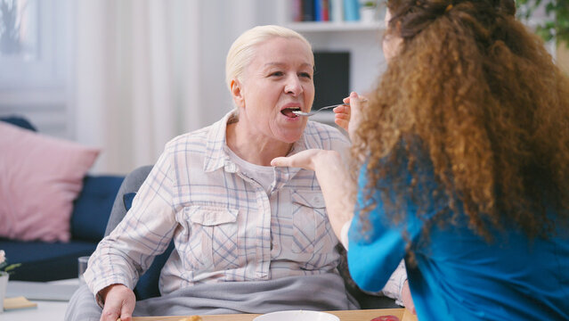 Caring nurse spoon feeding senior woman with disability, private home care services