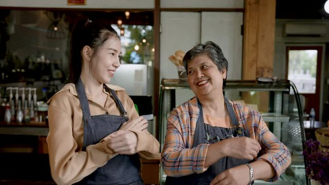 Senior mother with Asian daughter, standing in front checkout counter taking cafe, which is its own bakery, stood with their apron pockets in their pockets, raising their hands make hearts.