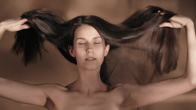 A woman demonstrates long silky, healthy and shiny hair. Seminude woman with hair flowing in the wind in the studio on a brown background with highlights. Keratin straightening. Slow motion.