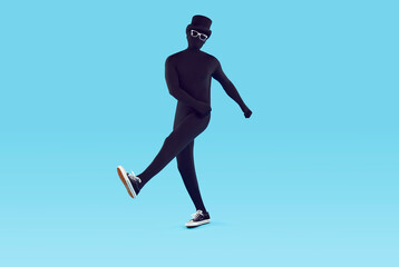 Fototapeta na wymiar Full length portrait of faceless unrecognizable person wearing black skinny bodysuit costume with hat and glasses in sneakers. Incognito funny man walking on a studio blue background.