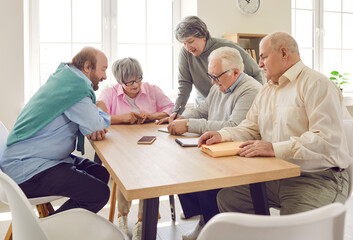Cheerful retired friends sitting around table at home. Positive senior men and women communicating, playing board game together in living room. Elderly people leisure activities, nursing home