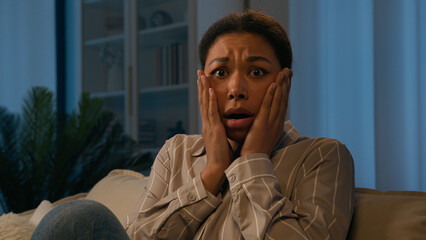 Scared African American woman frightened alone at evening home on couch shocked confused female...