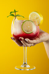 Cocktail rojo o bebida refrescante de zumos y alcohol. Tiene cítricos, menta y mucho hielo en una copa de cristal en forma de balón. Lo sujeta una mano femenina sobre un fondo amarillo.