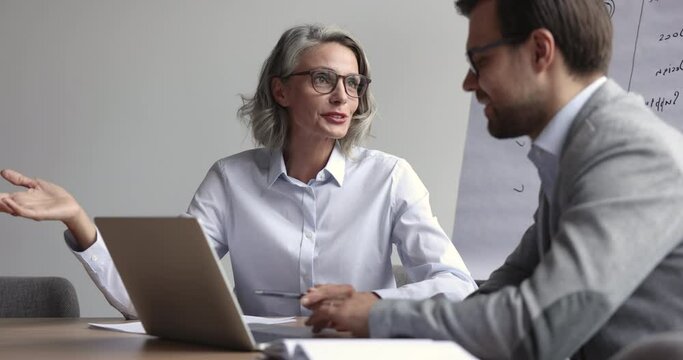 Confident beautiful mature business professional woman in glasses talking to younger male coworker at laptop, explaining work strategy, ideas for project, speaking with hand gesture for expression