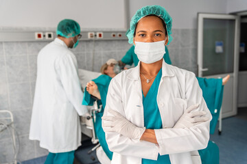 Photo of black beautiful midwife wearing surgical cap, mask and gloves standing with arms crossed while preparing for childbirth in operating theatre.