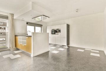 an empty living room with white walls and grey flooring, there is a small kitchen area in the corner