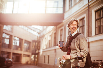 Caucasian middle aged man having takeaway coffee while pushing his bicycle and commuting to work