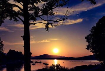 Zachód Słońca nad Jeziorem Żywieckim w sierpniu 2023, na jeziorze stoją zacumowane stateczki, niebo przyozdabiają białe obłoczki, wokół Słońca utworzyła się pomarańczowa poświata,