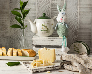 Hard cheese on a light wooden board. Behind sits a bunny and a beautiful teapot