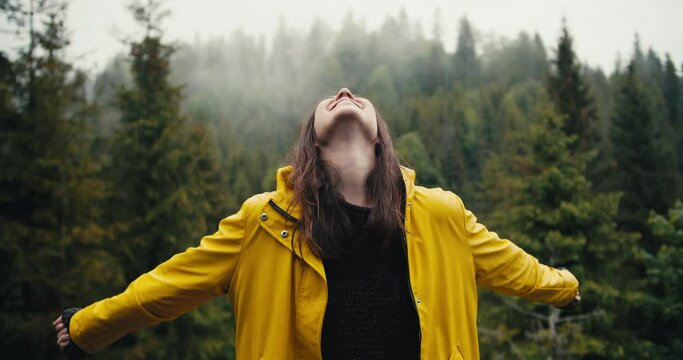 A happy girl in a yellow jacket stands against the background of a coniferous mountain forest, looks at the sky and rejoices in a little rain