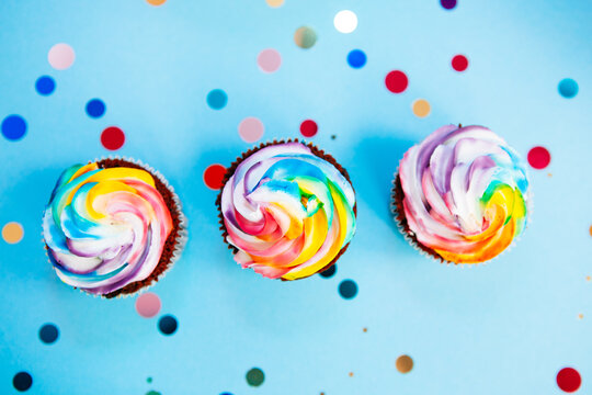 Three rainbow colors cupcake on blue background with glitter confetti from top view