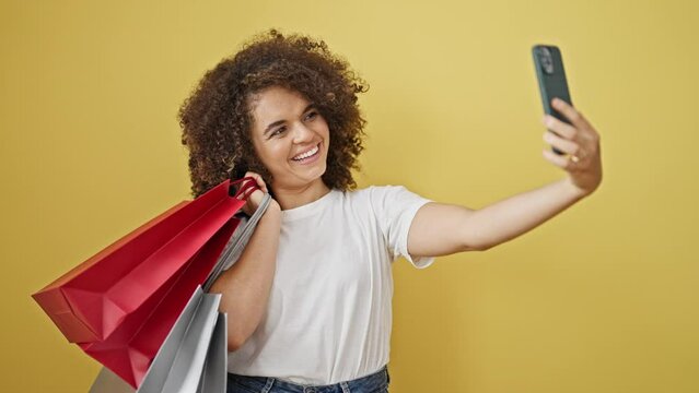 Young beautiful hispanic woman holding shopping bags make selfie by smartphone over isolated yellow background