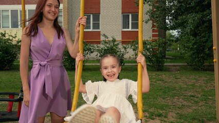 Careful mother plays swings with laughing daughter on children playground
