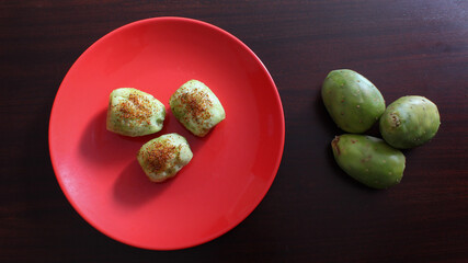 Plato rojo con tres tunas verdes sin cascara y tres tunas verdes con cascara sobre superficie de madera. 