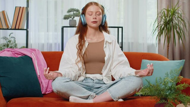 Teenager red hair girl breathes deeply with mudra gesture, eyes closed meditating with concentrated thoughts, peaceful mind. Child in wireless headphones relaxing listening music at home sits on sofa