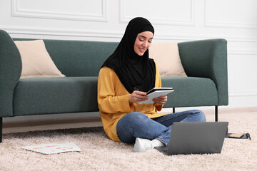 Muslim woman in hijab using laptop on floor near sofa indoors