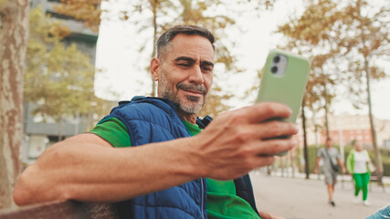 Close-up,happy mature man wearing casual clothes sitting on bench looking through information on smartphone