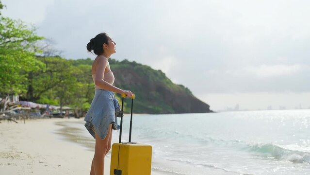 4K Young Asian woman tourist dragging a suitcase walking on tropical island beach at sunset. Attractive girl traveler relax and enjoy outdoor lifestyle travel ocean nature on summer holiday vacation.