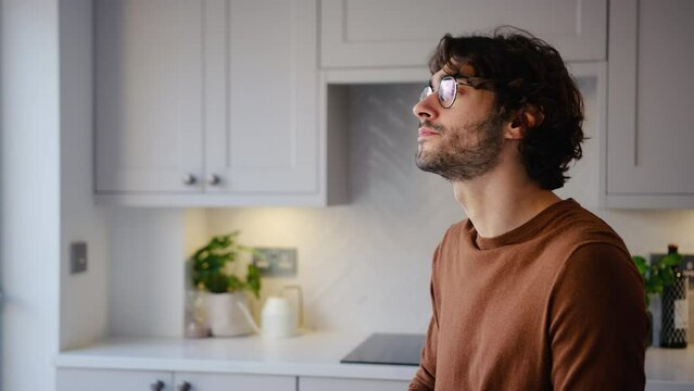 Side view portrait of smiling young man wearing glasses taking a break with cup of coffee sitting at kitchen counter at home with copy space on left side of frame - shot in slow motion