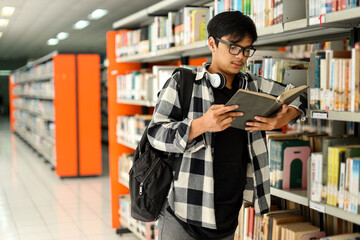 Young hipster Asian man student with headphones on neck, carrying backpack and reading a book inside campus library.