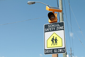 school safety zone drive slowly sign with illustration of people walking and flashing yellow light above with blue sky in background, black white and yellow