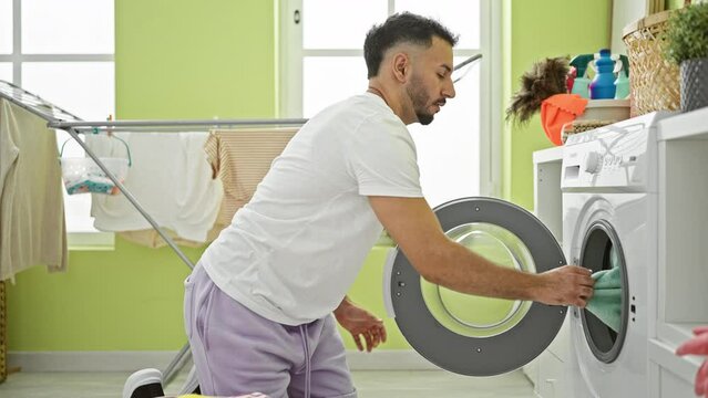 Young arab man washing clothes touching softener towel with face at laundry room