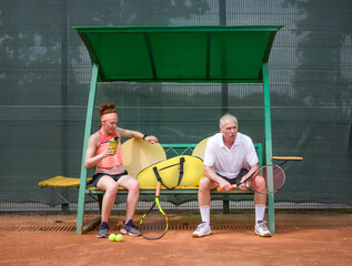 Tennis players are resting on the bench between sets. Mixed age. Sports family. Young woman and elderly man