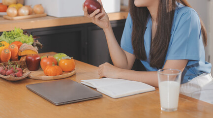 Obraz na płótnie Canvas A young woman with a beautiful face in a blue shirt with long hair eating fruit sitting inside the kitchen at home with a laptop and notebook for relaxation, Concept Vacation.