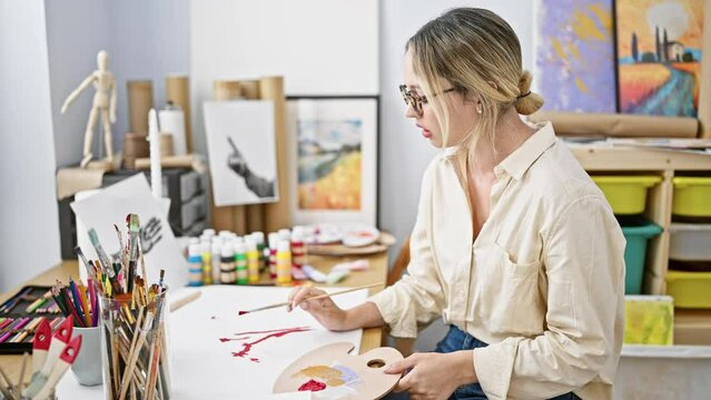 Young blonde woman artist drawing on paper taking glasses off tired at art studio