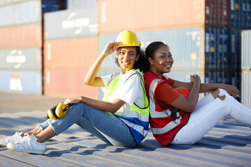 portrait young female African factory workers or engineers sitting back to back on container in warehouse storage