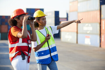 young female African factory workers or engineer using binoculars and pointing to something in containers warehouse storage