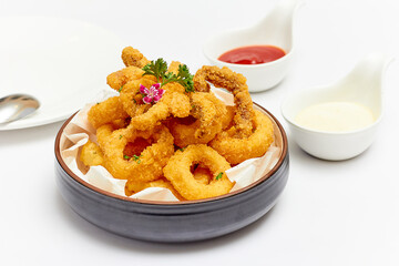 Calamari (deep-fried sliced squid rings) are beautifully decorated with flowers and parsley leaves and served with jagged French fries in a large bowl with sauce on white table. Popular Appetizer.