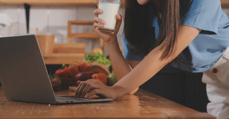 A young woman with a beautiful face in a blue shirt with long hair eating fruit sitting inside the kitchen at home with a laptop and notebook for relaxation, Concept Vacation.